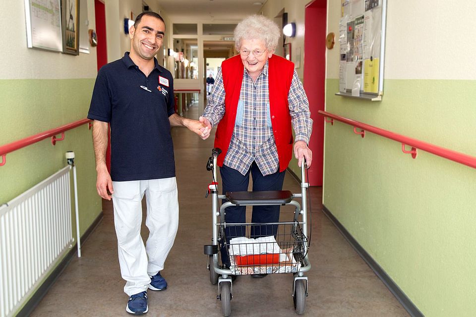 Eine Bewohnerin mit Rollator geht gemeinsam mit einem lächelnden Pfleger den Flur runter